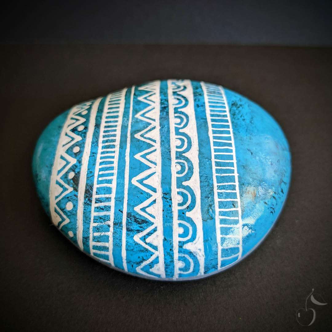 Ce galet peint à la main est conçu pour ressembler à une pierre précieuse, la turquoise. Il est orné d'un motif triangulaire typiquement mexicain pour un look unique et authentique.