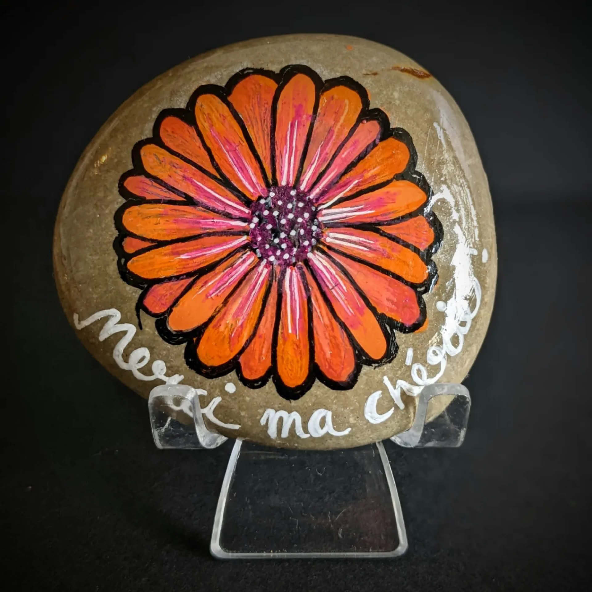 Galet naturel peint avec une fleur orange style marguerite. On peut lire sous la fleur "Merci ma chérie"