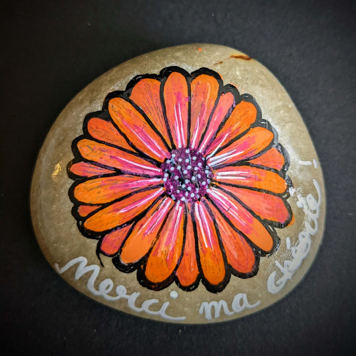 Galet naturel peint avec une fleur orange style marguerite. On peut lire sous la fleur "Merci ma chérie"
