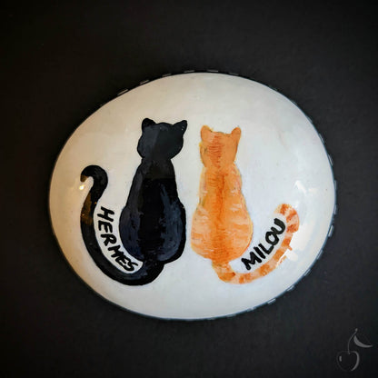 Galet peint blanc avec un dessin qui montre deux chat assis de dos. Un chat noir, Hermes et un chat roux, Milou