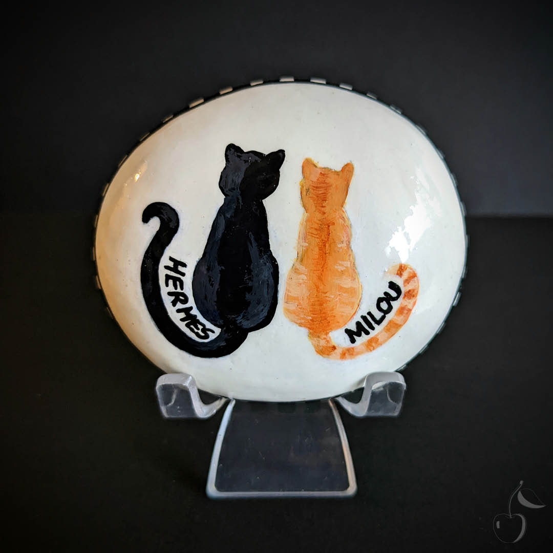 Galet peint blanc avec un dessin qui montre deux chat assis de dos. Un chat noir, Hermes et un chat roux, Milou