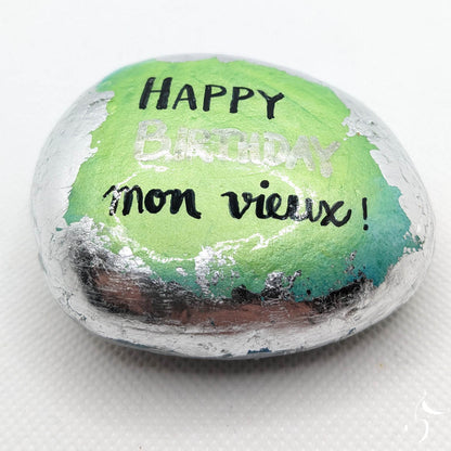 Galet peint de couleur vert/bleu aux bords en feuille argentée. Sur le galet est écrit : "Happy Birthday mon vieux".
