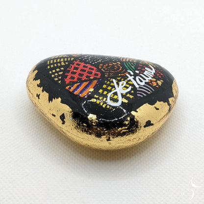 Galet en forme de coeur peint en noir avec formes géométriques et enveloppé dans une feuille dorée.