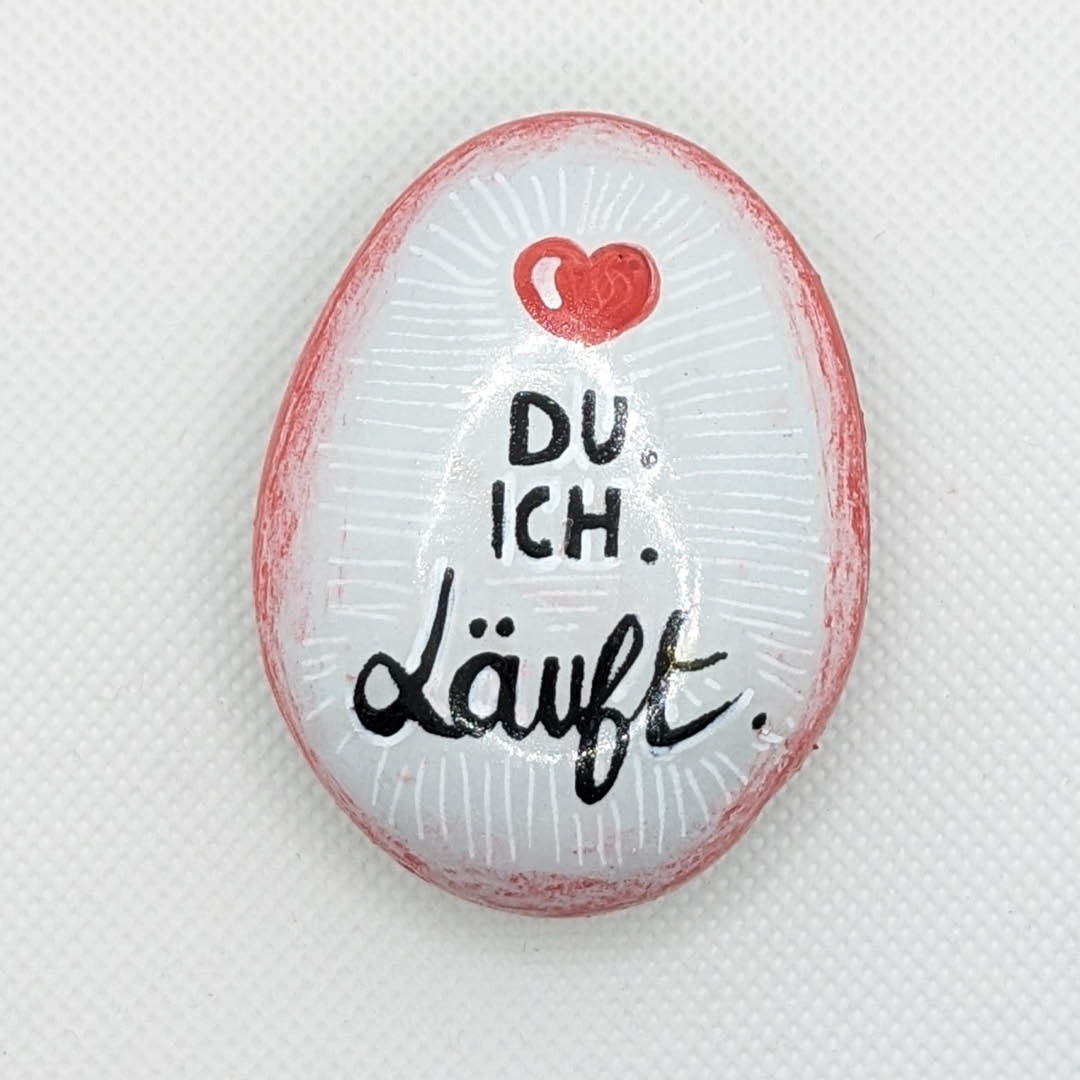 Galet peint en blanc avec un contour en dégradé rouge orné d'un coeur rouge. Inscription "DU. ICH. Läuft" en son centre. 