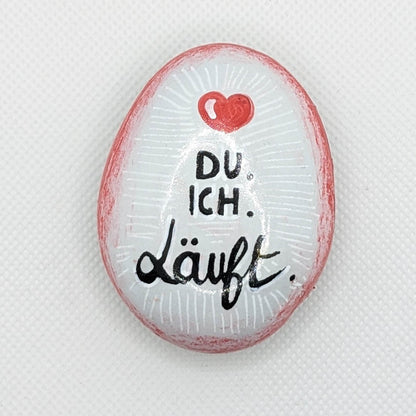 Galet peint en blanc avec un contour en dégradé rouge orné d'un coeur rouge. Inscription "DU. ICH. Läuft" en son centre. 