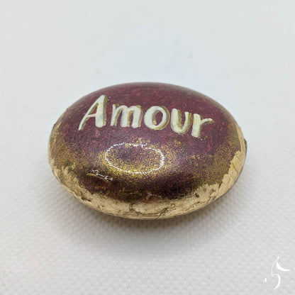 Galet peint en rose foncé métallisé. Bords en feuille dorée. Inscription "Amour" en blanc en son centre. Le tout recouvert d'un vernis brillant.